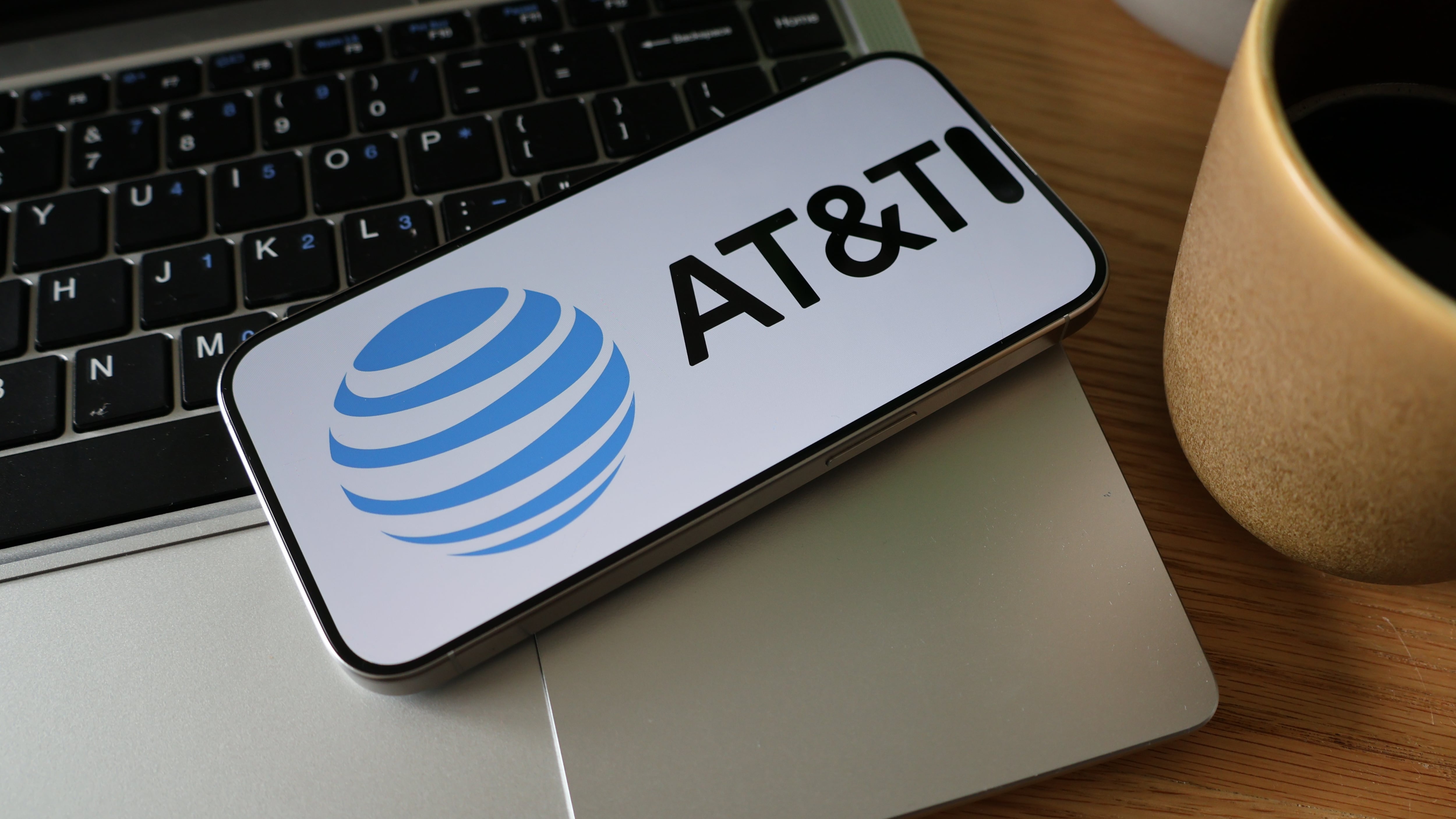 A smartphone displaying the AT&T logo rests on a laptop keyboard next to a coffee cup and a decorative pot